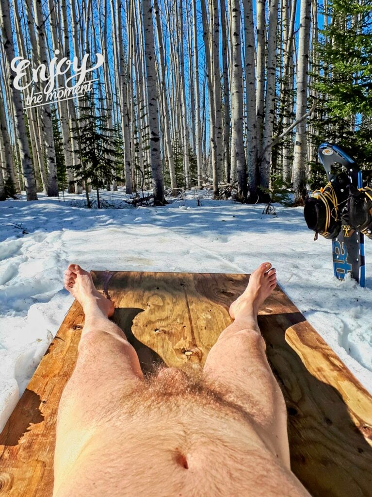 Shows a nude male laying on plywood on the snow with snowshoes to the background.